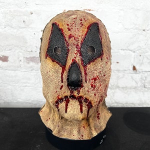 Puede incluir: Una máscara de saco de arpillera con un aspecto sangriento y desgastado. La máscara tiene dos agujeros negros para los ojos con malla detrás y una boca cosida.