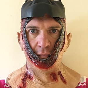 Może przedstawiać: Straszna maska ​​na Halloween z realistycznym efektem otwartej rany. Maska ma czarną g&oacute;rną część i dolną część w kolorze sk&oacute;ry z symulowaną krwią i odsłoniętą tkanką, tworząc przerażający wygląd.