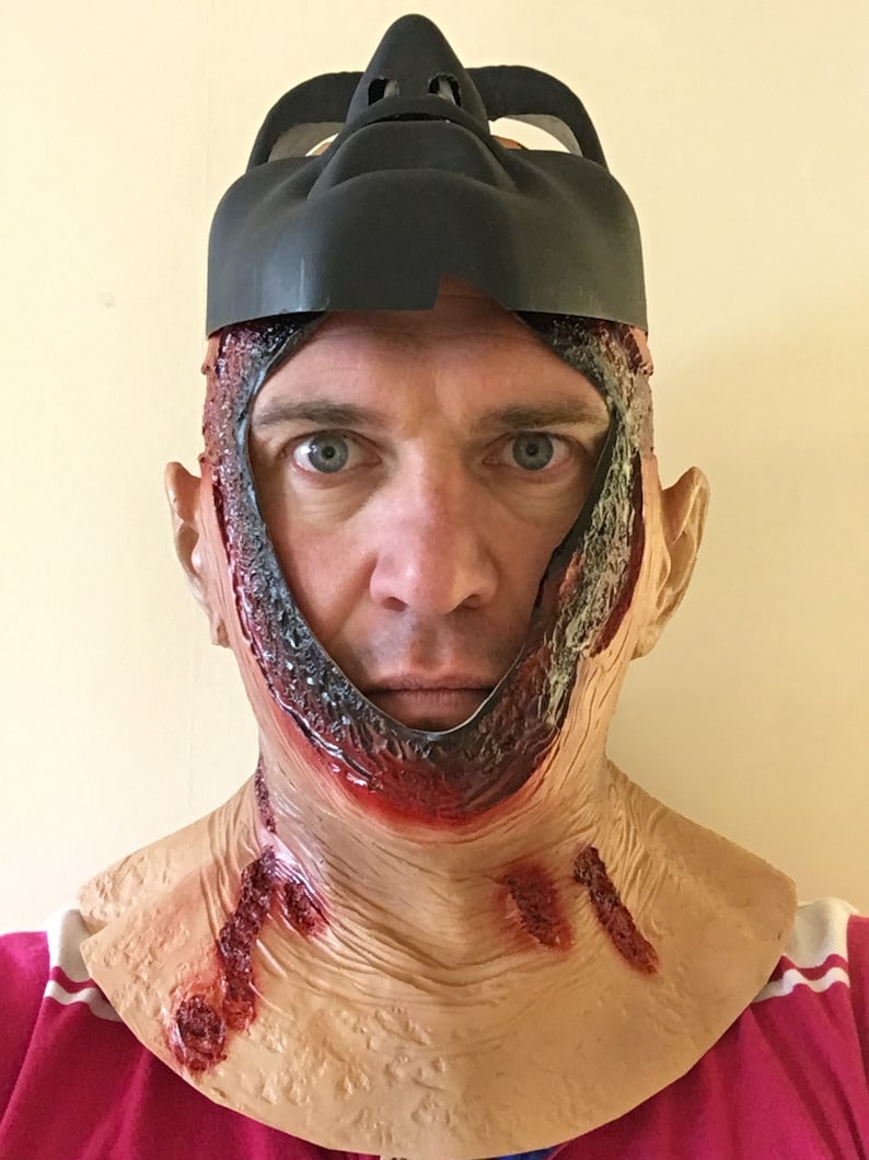 Może przedstawiać: Straszna maska ​​na Halloween z realistycznym efektem otwartej rany. Maska ma czarną g&oacute;rną część i dolną część w kolorze sk&oacute;ry z symulowaną krwią i odsłoniętą tkanką, tworząc przerażający wygląd.