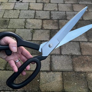 May include: A pair of large, black-handled scissors with silver blades. The scissors are open and being held by a hand.