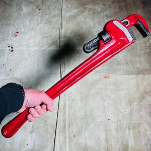 May include: A red pipe wrench with a silver handle. The wrench is held by a hand.