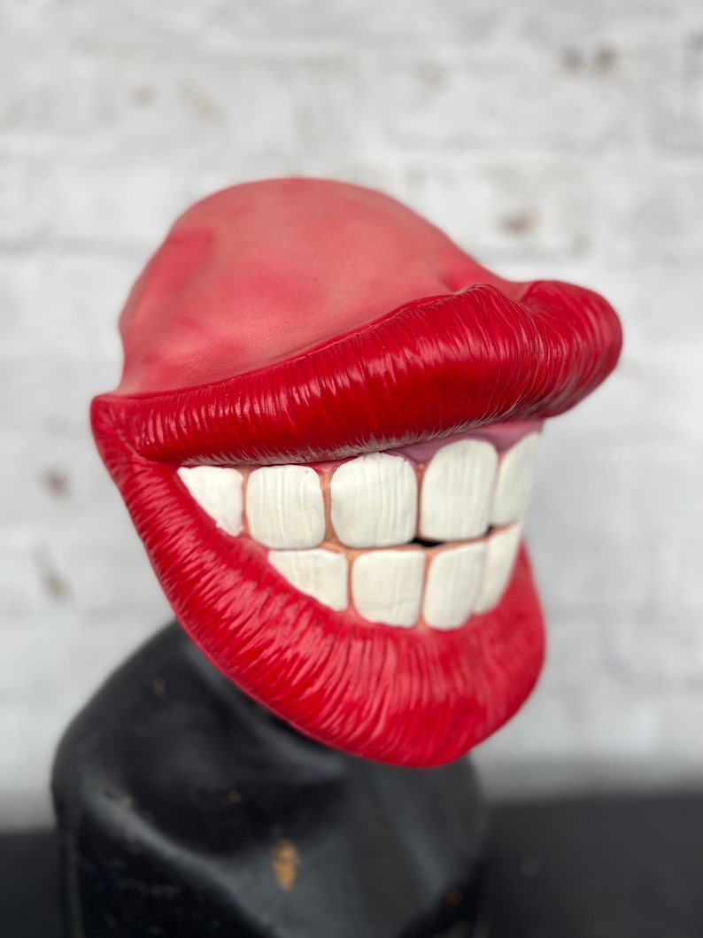 Puede incluir: Una m&aacute;scara de pl&aacute;stico roja y blanca con una gran sonrisa de dientes y labios exagerados. La m&aacute;scara est&aacute; dise&ntilde;ada para parecer un personaje de dibujos animados.