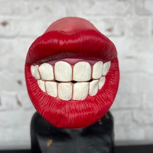 Puede incluir: Una boca grande, roja y esculpida con dientes blancos, sonriendo ampliamente. La boca est&aacute; hecha de un material liso y brillante y parece ser un objeto decorativo.