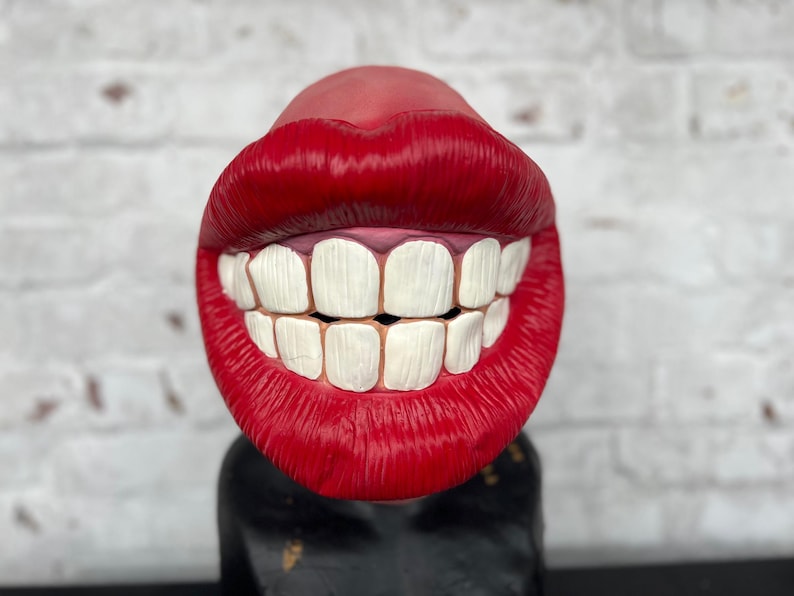 Puede incluir: Una boca grande, roja y esculpida con dientes blancos, sonriendo ampliamente. La boca est&aacute; hecha de un material liso y brillante y parece ser un objeto decorativo.