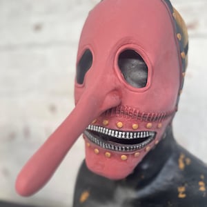 May include: A red mask with a long nose and a zipper mouth. The mask has eye holes and is adorned with gold-colored rivets. The top of the mask is black with gold accents. The mask is on a black surface.
