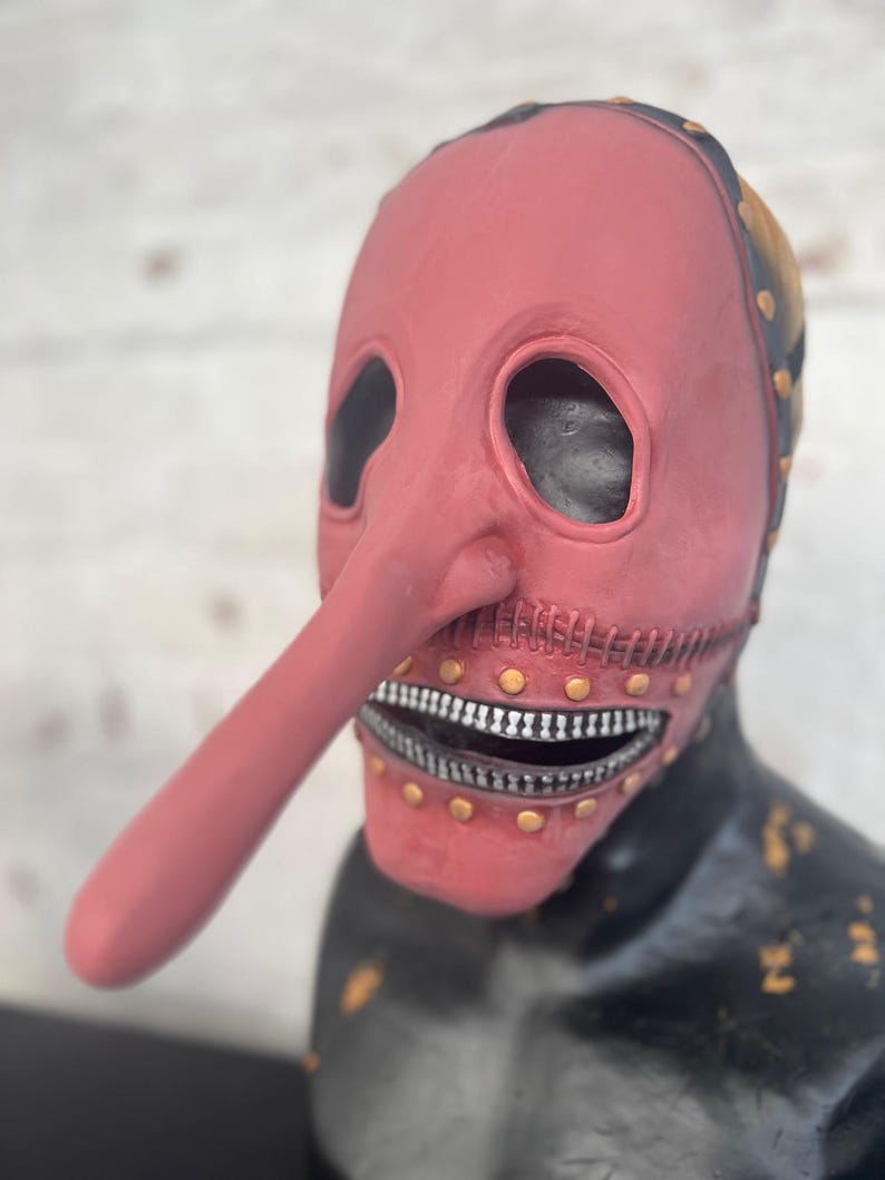 May include: A red mask with a long nose and a zipper mouth. The mask has eye holes and is adorned with gold-colored rivets. The top of the mask is black with gold accents. The mask is on a black surface.