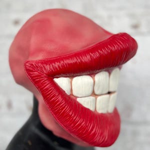 Puede incluir: Una m&aacute;scara de goma roja con una gran sonrisa exagerada. La m&aacute;scara tiene dientes blancos y labios rojos.