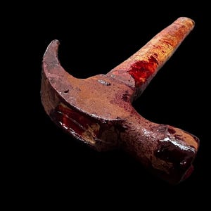 Pu&ograve; includere: Un martello arrugginito con un manico di legno ricoperto di sangue. Il martello &egrave; sdraiato di lato con la testa rivolta verso lo spettatore.