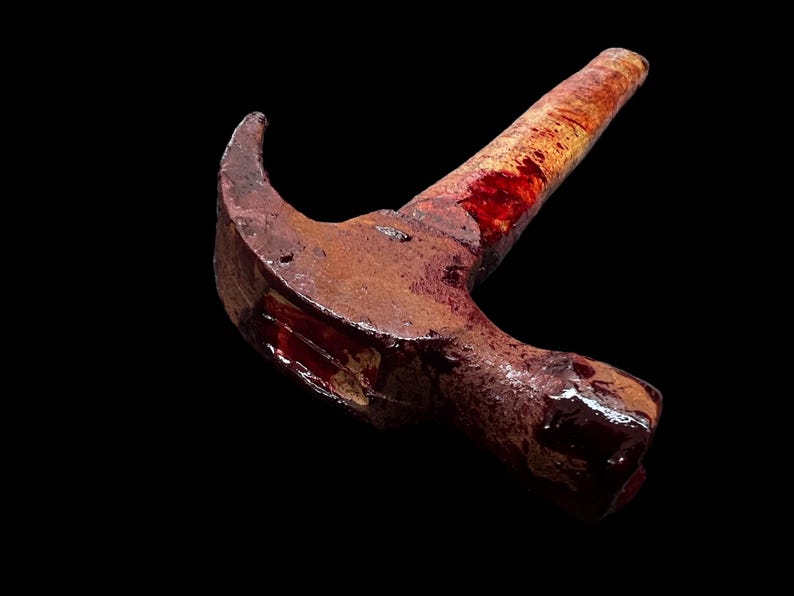 Pu&ograve; includere: Un martello arrugginito con un manico di legno ricoperto di sangue. Il martello &egrave; sdraiato di lato con la testa rivolta verso lo spettatore.