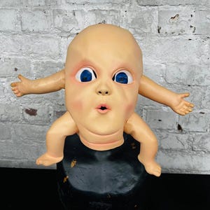 May include: A vintage, grotesque baby doll sculpture with a peach-colored head, large blue eyes, and outstretched arms. The doll is mounted on a black, rounded base. The background is a white brick wall.