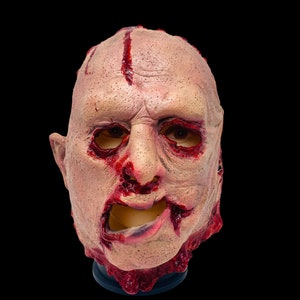 May include: A horrifying flesh-toned mask with realistic details, featuring open eye sockets and a gaping mouth. Red, blood-like paint is splattered across the forehead, nose, and mouth, creating a gruesome effect. The mask is designed for Halloween or costume use.