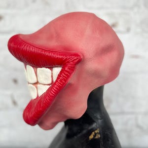 Puede incluir: Una m&aacute;scara de pl&aacute;stico roja y blanca con una gran sonrisa exagerada. La m&aacute;scara tiene labios rojos y dientes blancos.