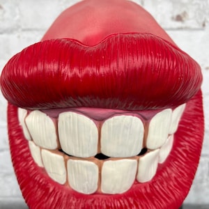 Puede incluir: Una m&aacute;scara de pl&aacute;stico roja con una gran sonrisa de dientes y labios rojos brillantes. La m&aacute;scara est&aacute; dise&ntilde;ada para parecer una boca gigante.