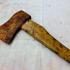 K&ouml;nnte beinhalten: Eine rostige Axt mit einem gelben Holzstiel. Die Axt liegt auf einer wei&szlig;en Oberfl&auml;che.