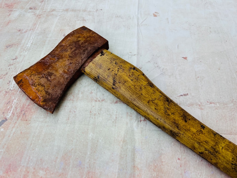 K&ouml;nnte beinhalten: Eine rostige Axt mit einem gelben Holzstiel. Die Axt liegt auf einer wei&szlig;en Oberfl&auml;che.