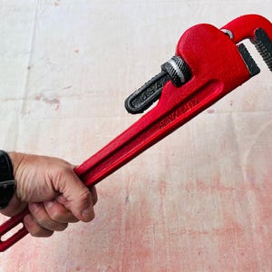 May include: A red pipe wrench with a black handle and a silver adjustment knob. The wrench is held in a hand.