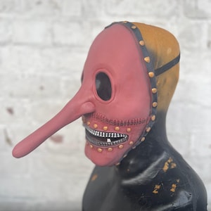 May include: A red plague doctor mask with a long, curved beak and large eye openings. The mask has a zipper-like mouth and is attached to a black head covering with gold rivets. The mask is displayed on a black mannequin head.