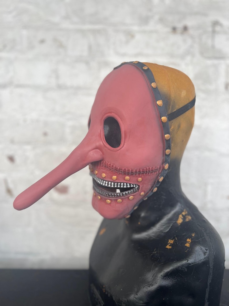 May include: A red plague doctor mask with a long, curved beak and large eye openings. The mask has a zipper-like mouth and is attached to a black head covering with gold rivets. The mask is displayed on a black mannequin head.