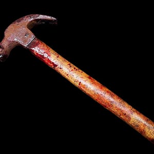 Pu&ograve; includere: Un martello arrugginito con un manico di legno macchiato di rosso. Il martello &egrave; isolato su uno sfondo nero.