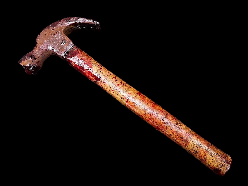 Pu&ograve; includere: Un martello arrugginito con un manico di legno macchiato di rosso. Il martello &egrave; isolato su uno sfondo nero.
