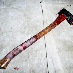 Puede incluir: Un hacha ensangrentada con una cabeza negra y un mango de madera manchado de pintura roja y amarilla.