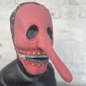 May include: A red plague doctor mask with a long, curved nose and a zipper mouth. The mask has black trim with gold rivets and eye holes. The mask is displayed on a black bust.