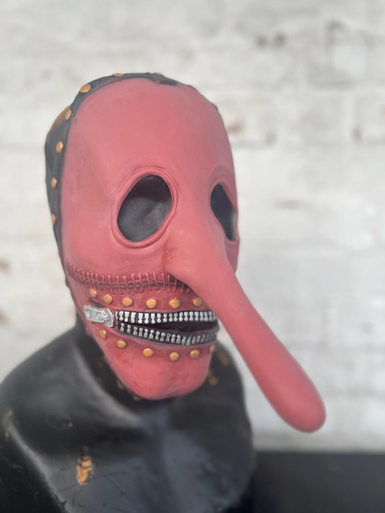 May include: A red plague doctor mask with a long, curved nose and a zipper mouth. The mask has black trim with gold rivets and eye holes. The mask is displayed on a black bust.
