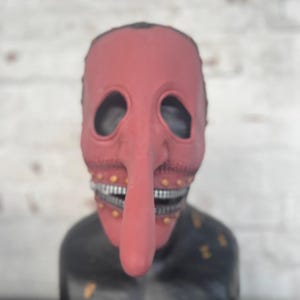 May include: A red mask with a long nose and a stitched mouth. The mask has eye holes and a black base with gold accents. The mask is displayed against a white brick wall.