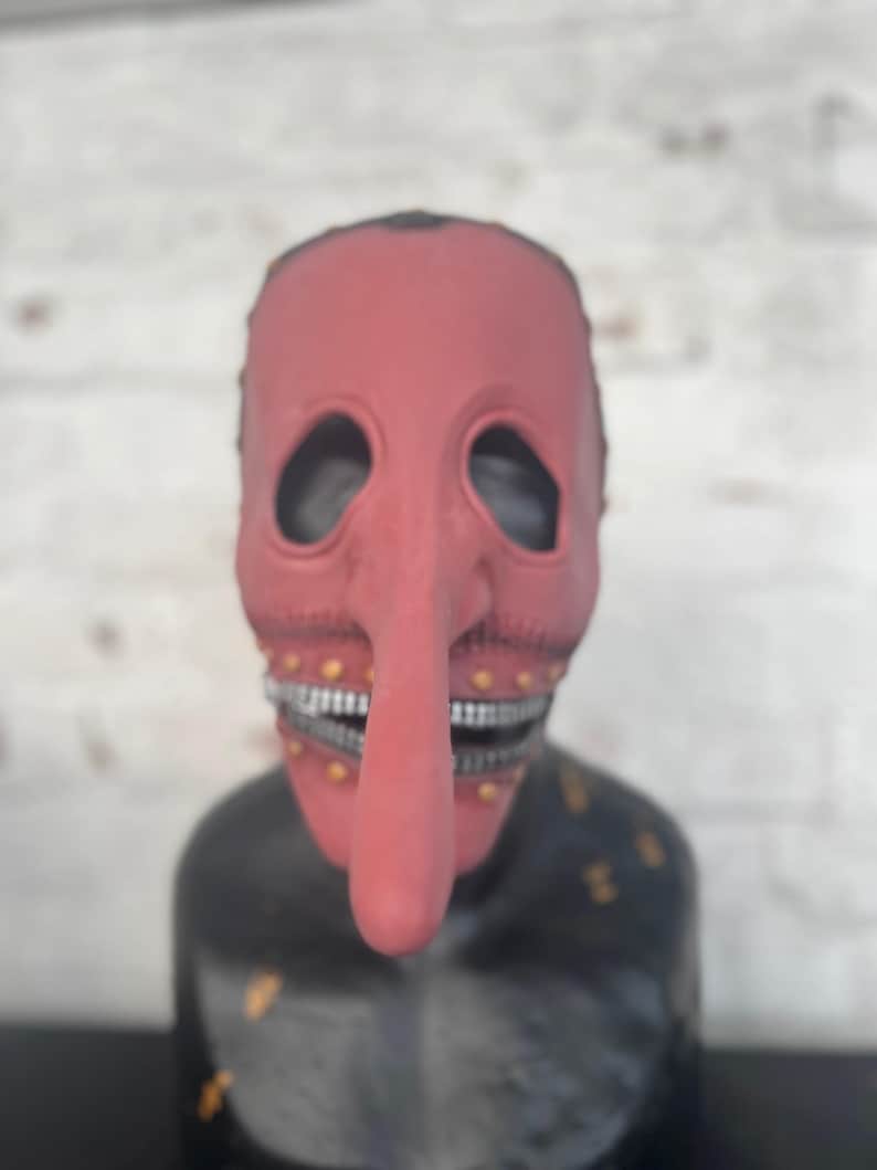 May include: A red mask with a long nose and a stitched mouth. The mask has eye holes and a black base with gold accents. The mask is displayed against a white brick wall.