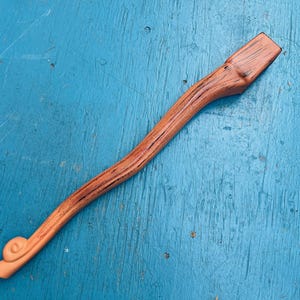 May include: A wooden tool with a curved handle and a rectangular head. The wood has a natural grain and a warm, reddish-brown colour. The handle features a decorative spiral at the end.