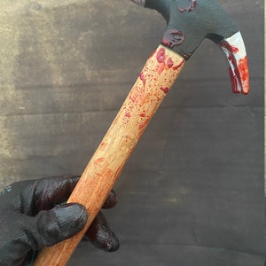 May include: A black and red hammer with a wooden handle covered in red paint splatters. The hammer is held in a black gloved hand.