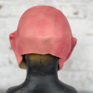 Puede incluir: Una m&aacute;scara de l&aacute;tex rosa con una superficie lisa. La m&aacute;scara se lleva puesta en una cabeza de maniqu&iacute; negra.