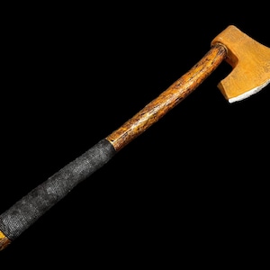 Puede incluir: Un hacha con mango de madera, mango marrón y negro y hoja dorada. El hacha está aislada sobre un fondo negro.