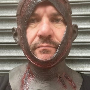 Może przedstawiać: Realistyczna maska ​​na Halloween z ciemnobrązowym nakryciem głowy poplamionym krwią i szarą częścią szyi. Maska ma duży otw&oacute;r na twarz i szczeg&oacute;łowe nacięcia, tworząc przerażający efekt. Idealna na straszny kostium.