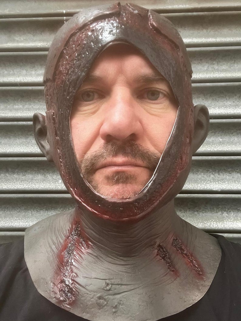 Może przedstawiać: Realistyczna maska ​​na Halloween z ciemnobrązowym nakryciem głowy poplamionym krwią i szarą częścią szyi. Maska ma duży otw&oacute;r na twarz i szczeg&oacute;łowe nacięcia, tworząc przerażający efekt. Idealna na straszny kostium.
