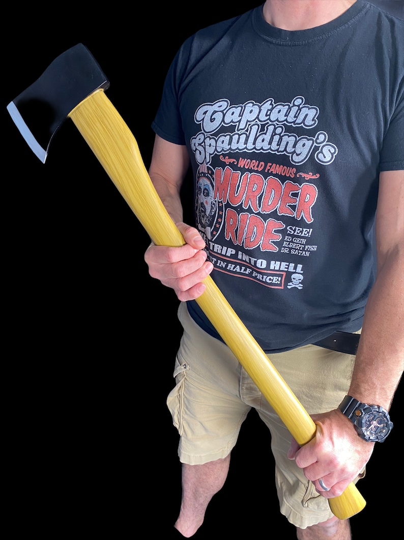 K&ouml;nnte beinhalten: Eine Person h&auml;lt eine Axt mit schwarzem Griff und einem Holzstiel. Die Axt wird in bedrohlicher Weise gehalten. Die Person tr&auml;gt ein schwarzes T-Shirt mit dem Aufdruck "Captain Spaulding's World Famous Murder Ride". Der Text "See! Ed Gein, Elbert Fish, Dr. Satan Trip Into Hell 1/2 Price!" ist ebenfalls auf dem Shirt aufgedruckt.