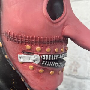 May include: A red plague doctor mask with a long, curved beak and large eye openings. The mask features a zipper mouth design with silver teeth and is adorned with gold-colored rivets. The mask has a stitched seam across the mouth.