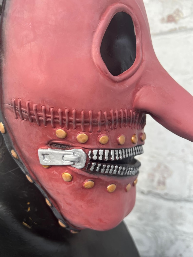 May include: A red plague doctor mask with a long, curved beak and large eye openings. The mask features a zipper mouth design with silver teeth and is adorned with gold-colored rivets. The mask has a stitched seam across the mouth.