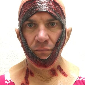 Może przedstawiać: Straszna maska ​​na Halloween o realistycznym, krwawym designie. Maska ma rozdartej sk&oacute;rze głowy i odsłoniętym ciele, z krwistoczerwonymi akcentami i rozdziawioną raną wok&oacute;ł twarzy i szyi. Idealna na kostium horror.