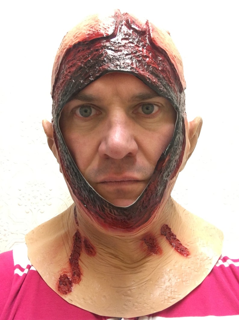Może przedstawiać: Straszna maska ​​na Halloween o realistycznym, krwawym designie. Maska ma rozdartej sk&oacute;rze głowy i odsłoniętym ciele, z krwistoczerwonymi akcentami i rozdziawioną raną wok&oacute;ł twarzy i szyi. Idealna na kostium horror.