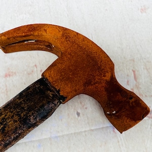 Pu&ograve; includere: Un martello arrugginito con un manico di legno. La testa del martello &egrave; di colore arancione brillante con macchie di ruggine marroni. Il manico &egrave; marrone scuro e ha una finitura usurata.