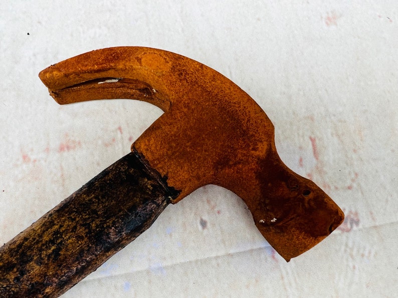 Pu&ograve; includere: Un martello arrugginito con un manico di legno. La testa del martello &egrave; di colore arancione brillante con macchie di ruggine marroni. Il manico &egrave; marrone scuro e ha una finitura usurata.