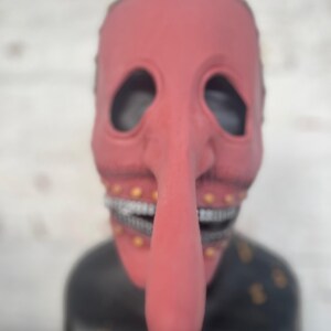 May include: A red mask with a long nose and large eye holes. The mask has a wide, toothy grin and small gold accents. The mask is set against a blurred background.