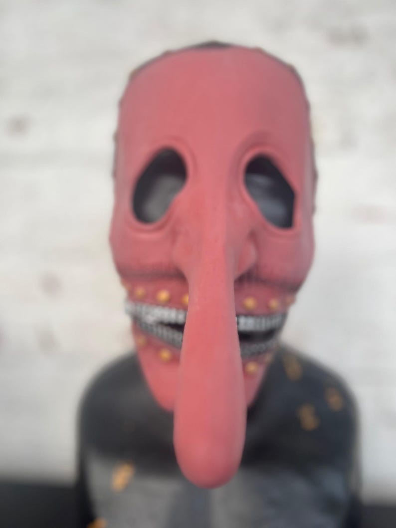 May include: A red mask with a long nose and large eye holes. The mask has a wide, toothy grin and small gold accents. The mask is set against a blurred background.