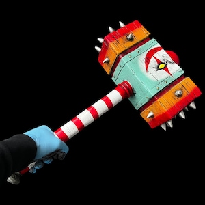 Könnte beinhalten: Ein Holzhammer mit einem rot-weiß-blau gestreiften Stiel. Der Kopf ist mit Spikes besetzt und in blau, weiß und orange bemalt, mit einem schwarzen Auge und einer weißen Pupille in der Mitte.