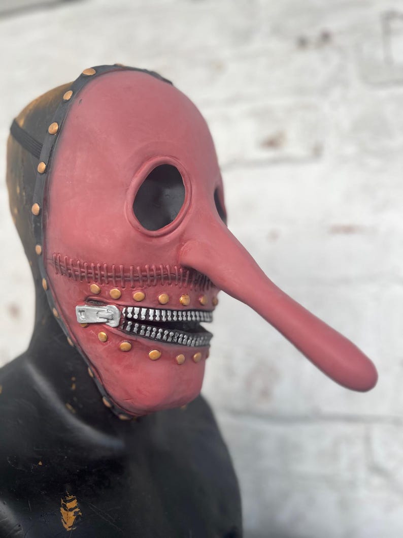May include: A red plague doctor mask with a long, curved nose and eye holes. The mask has a zipper for a mouth and is adorned with gold rivets. The top of the mask is black with gold rivets and a black strap.