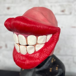 Puede incluir: Una m&aacute;scara de pl&aacute;stico roja y blanca con una gran sonrisa de dientes y labios exagerados. La m&aacute;scara est&aacute; dise&ntilde;ada para parecerse a la boca de una persona.