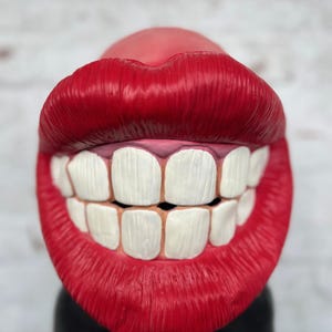 Puede incluir: Una boca grande de pl&aacute;stico rojo con dientes blancos sonr&iacute;e ampliamente. La boca es un art&iacute;culo novedoso y podr&iacute;a usarse como accesorio o decoraci&oacute;n.