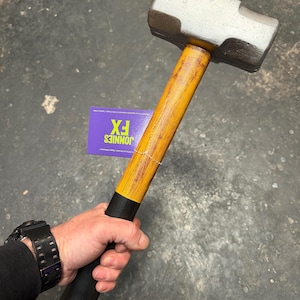 Può includere: Una mazza con testa color argento metallizzato e manico in legno giallo con un'impugnatura nera alla base. Un'etichetta rettangolare viola attaccata al manico mostra il testo JONNIES FX in lettere gialle su uno sfondo viola.