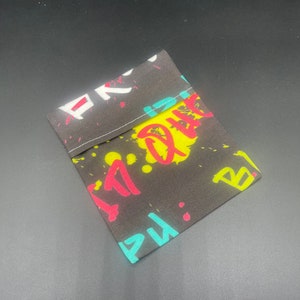 Puede incluir: Una pequeña bolsa negra con cierre de cremallera. La bolsa está hecha de tela y tiene un estampado de estilo graffiti colorido con salpicaduras de pintura amarilla, roja, azul y blanca y letras.